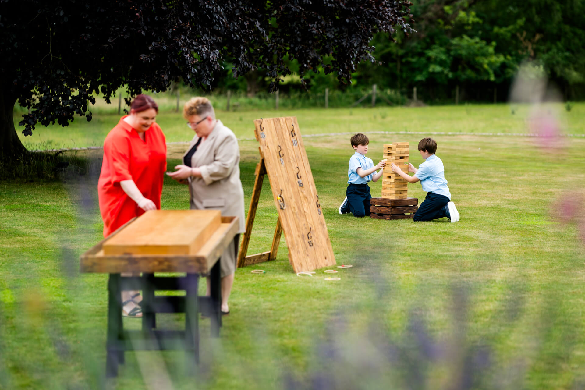 Two women stand by a wooden table, while two boys kneel on grass playing a large Jenga-like game in a park or garden setting.