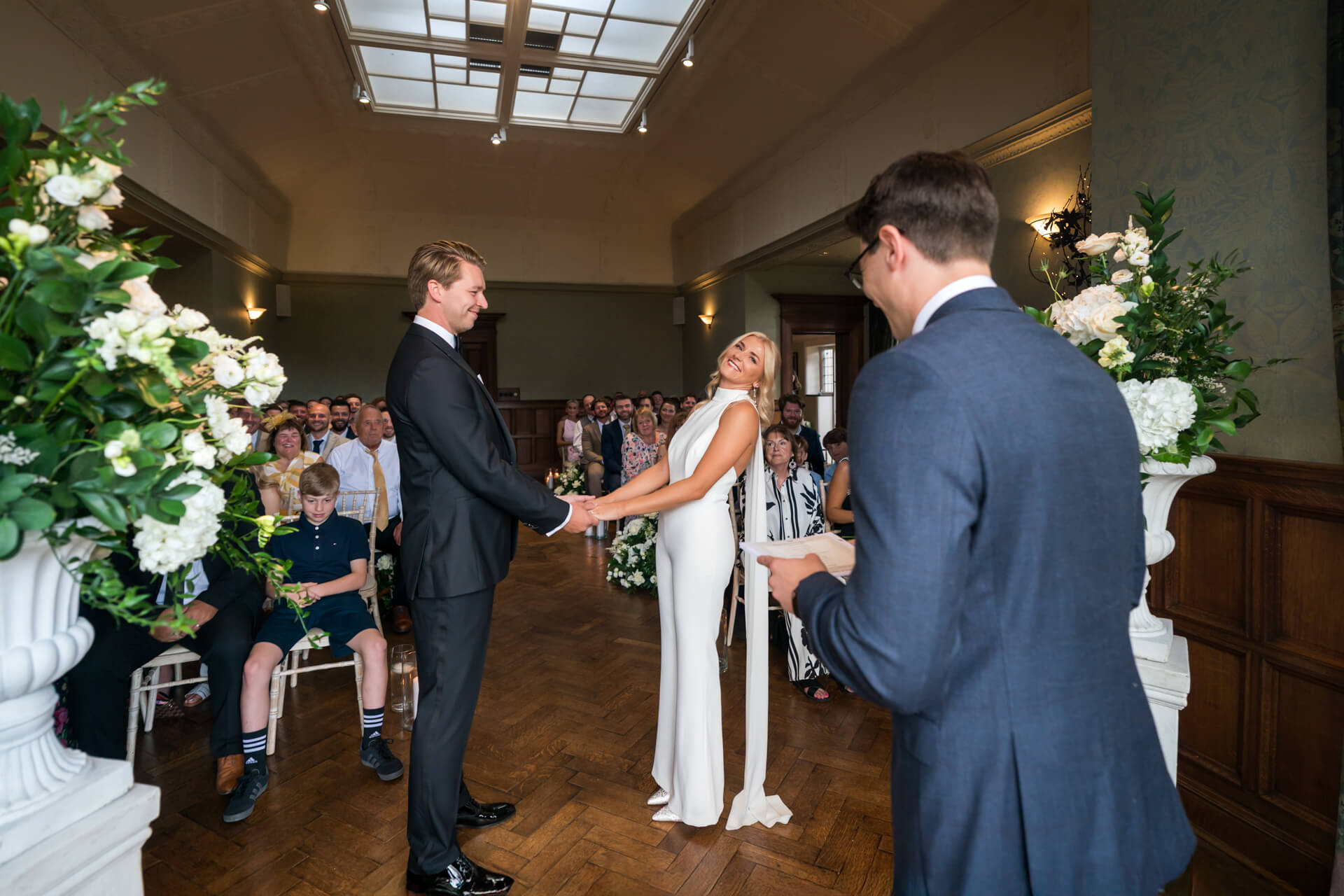 A couple stands holding hands at the altar during a wedding ceremony, facing an officiant, with guests seated and watching in the background.