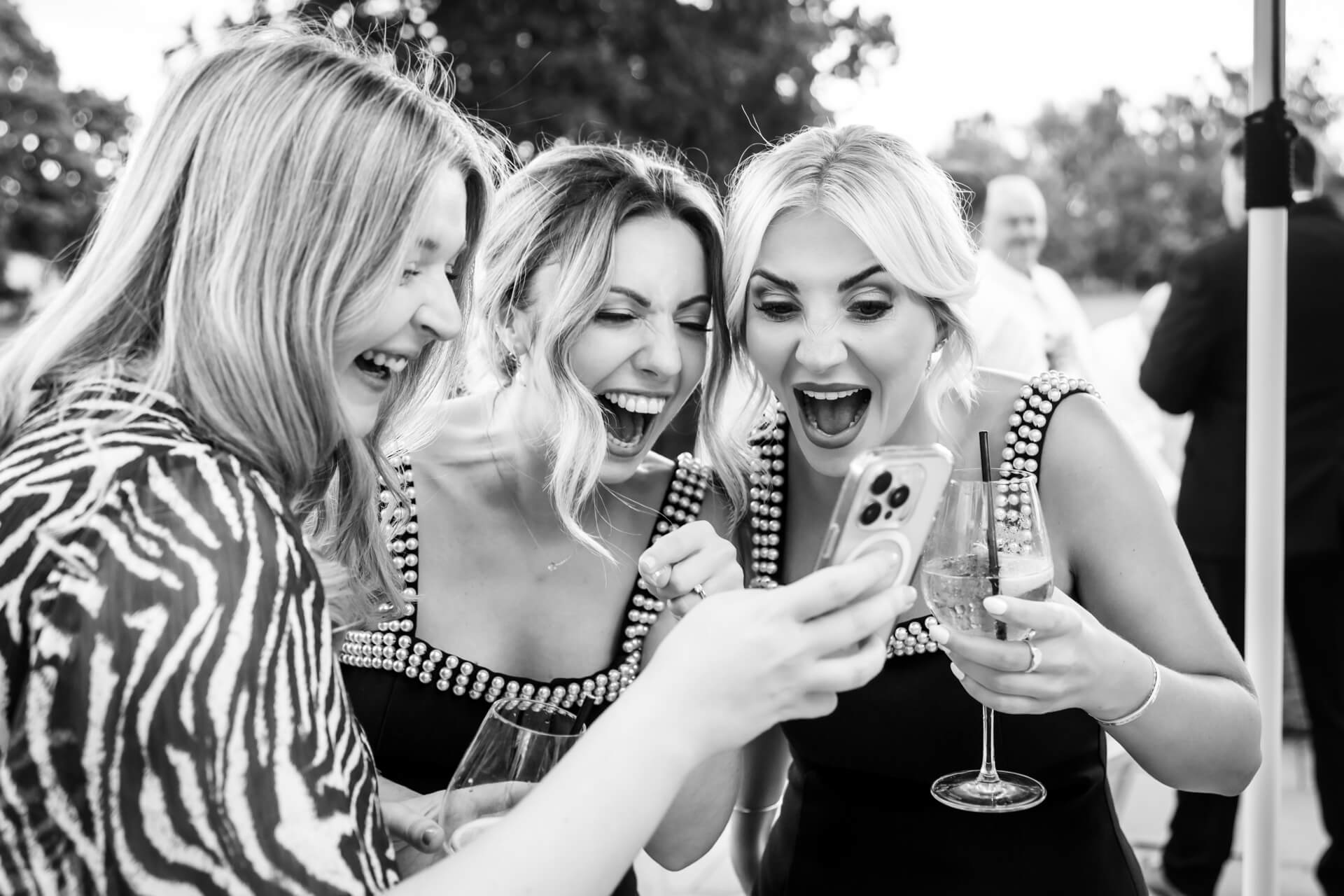 Three women dressed for a wedding laugh excitedly whilst looking at a mobile, one holding a glass of wine. The image is in black and white.