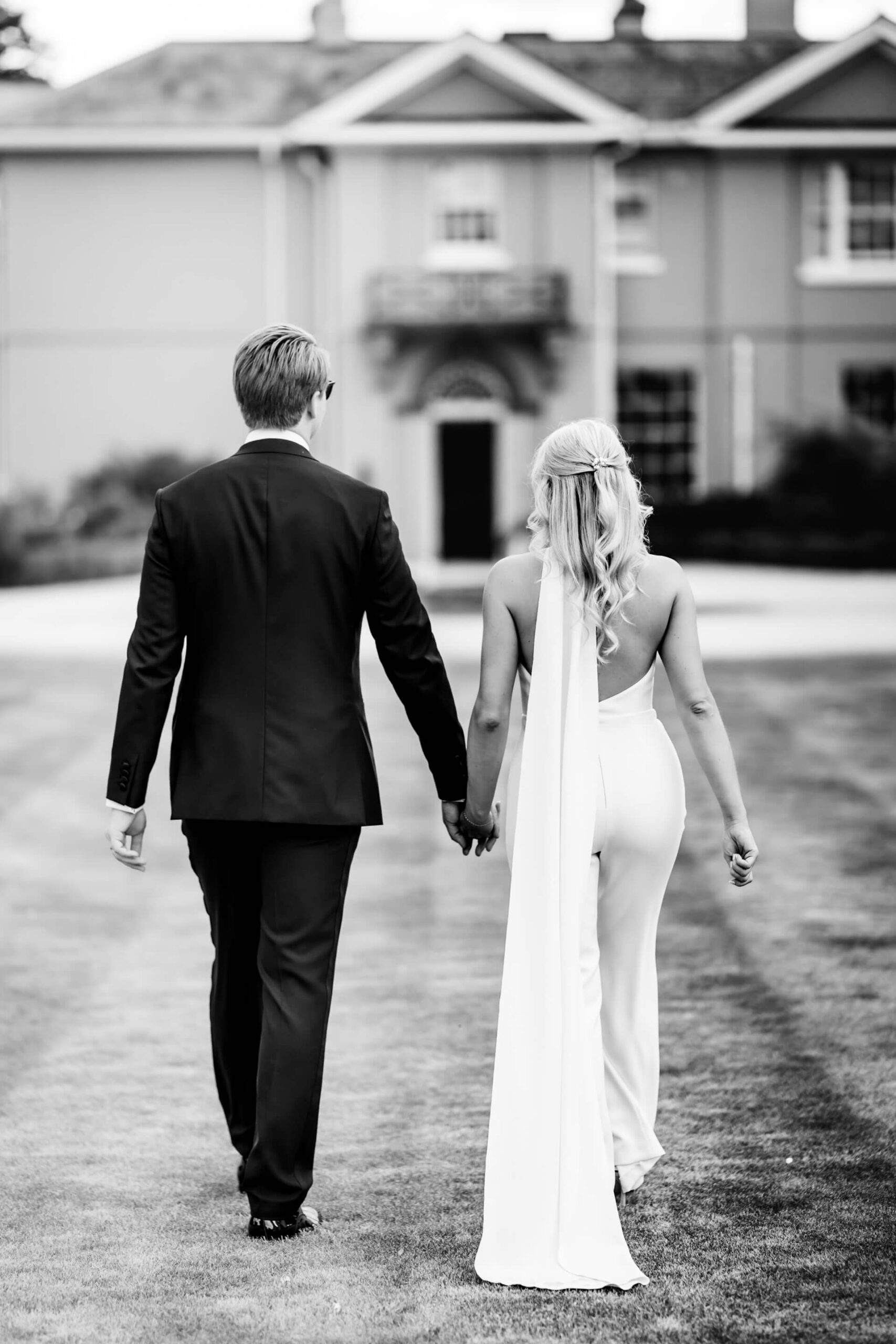 A wedding couple walk hand in hand towards a large house, photographed from behind in black and white.