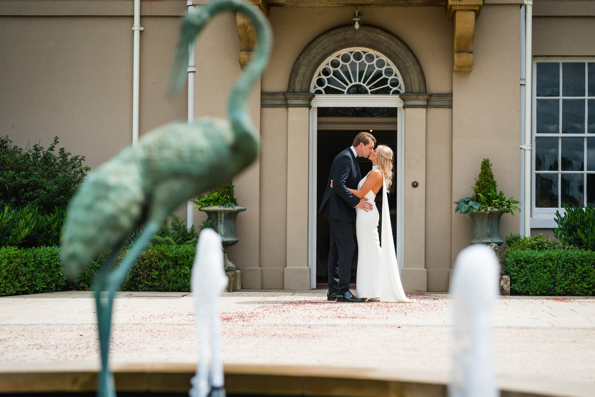A bride and groom kiss in front of the Burtonfields Hall entrance, with a large bird statue and fountains in the foreground.