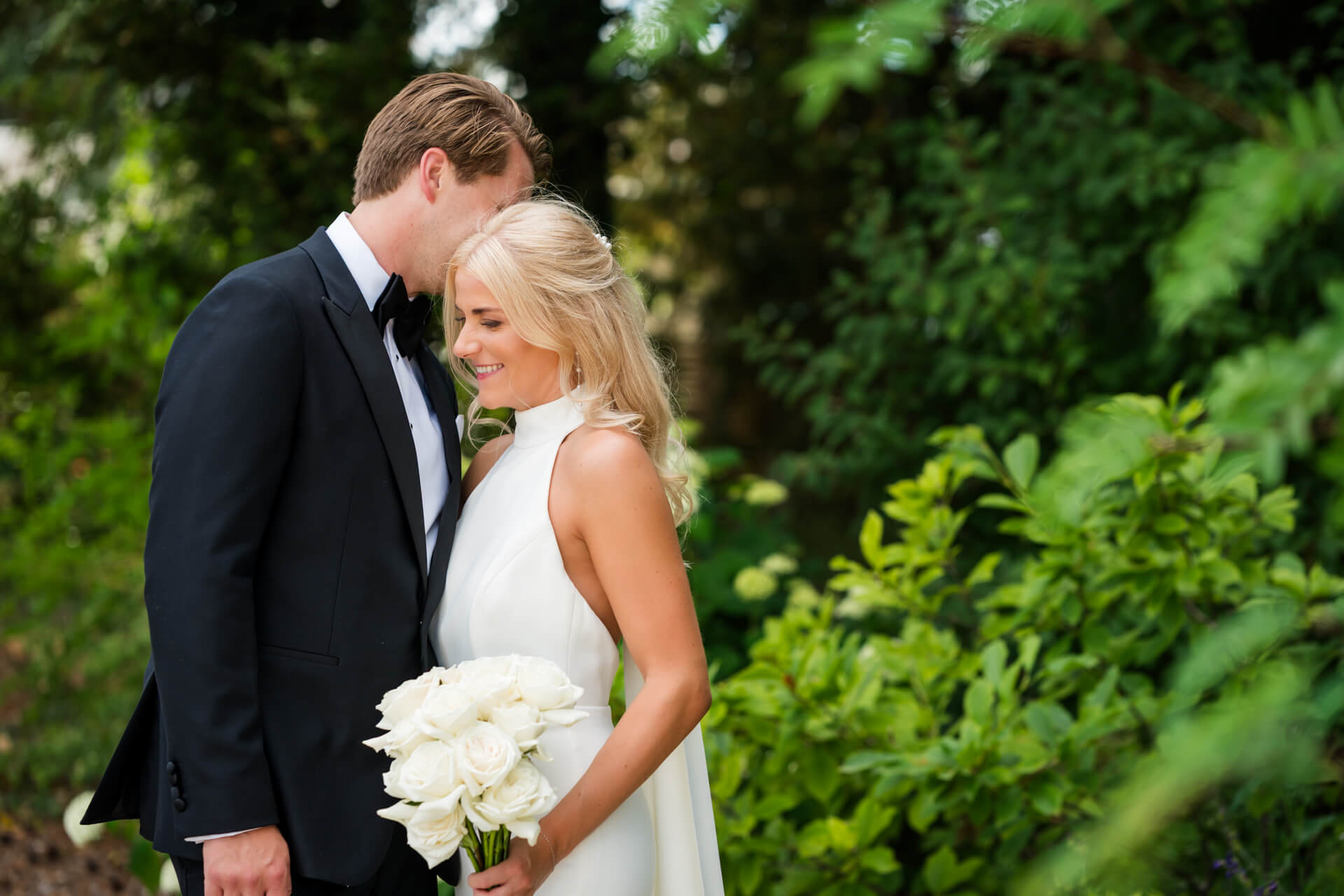 A bride in a white dress holding a bouquet of white roses stands beside a groom in a black suit, both smiling, surrounded by greenery.