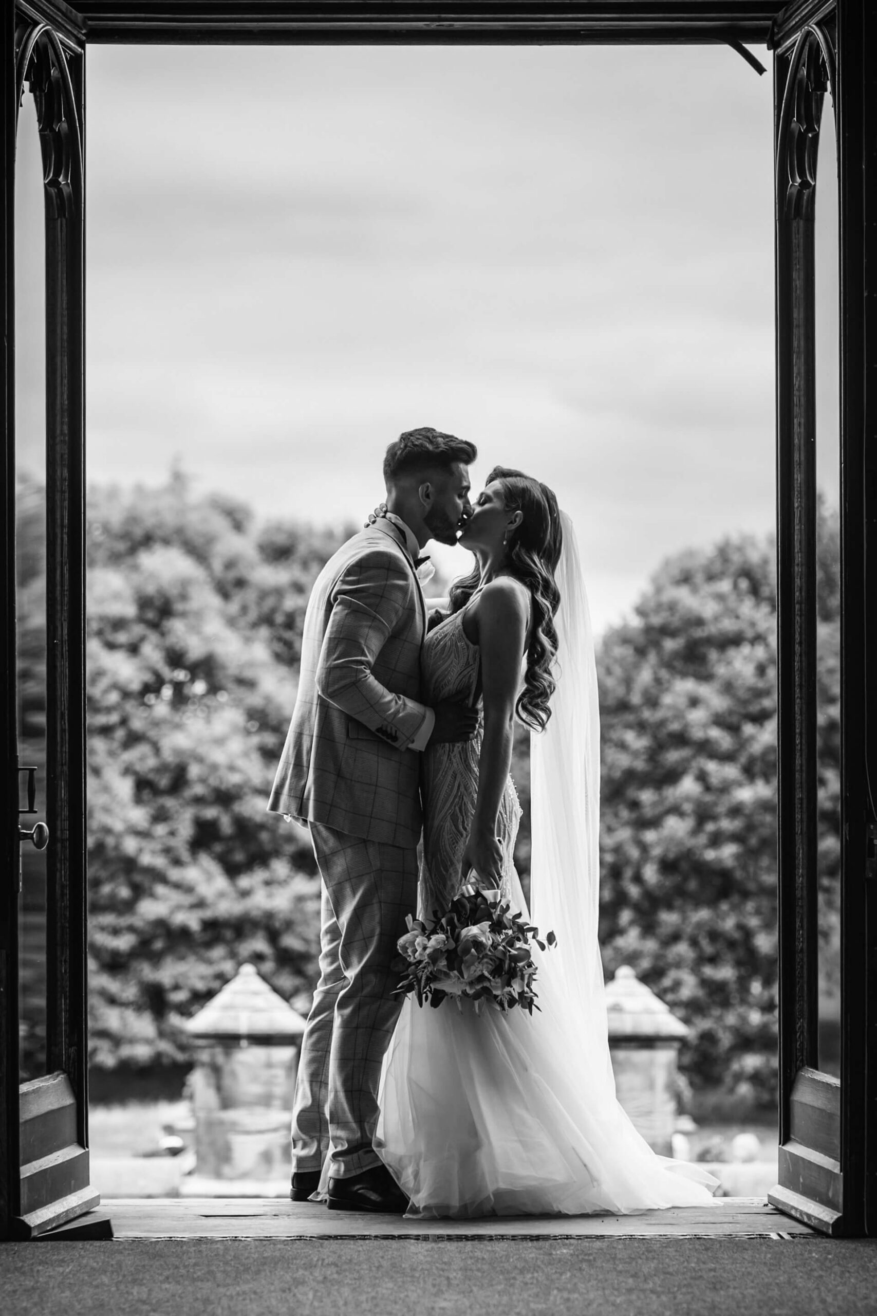 Newlywed couple kissing in doorway, holding bouquet.