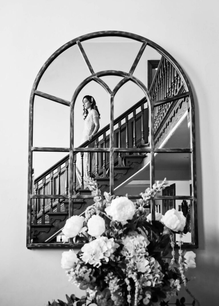A woman stands on a staircase, reflected in a large arched mirror above a floral arrangement in a black and white photo.
