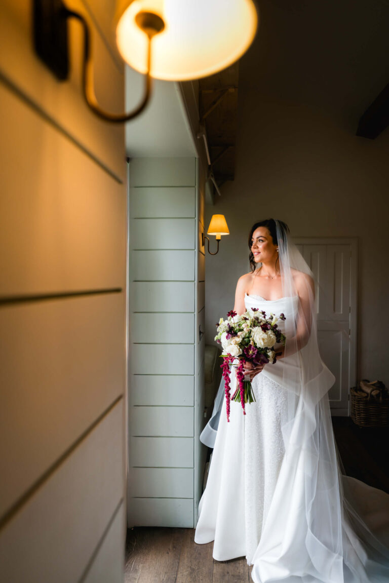 Bride in white dress holding floral bouquet indoors.