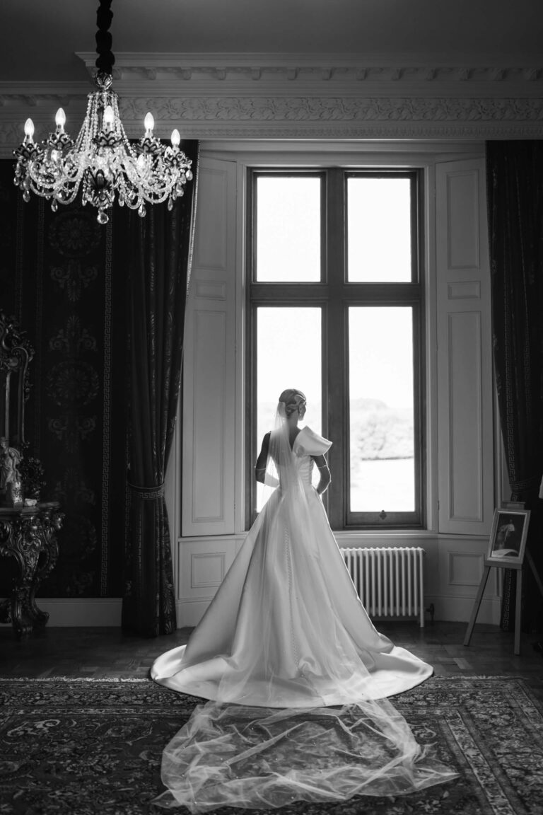 Bride in elegant dress by large window, chandelier above.
