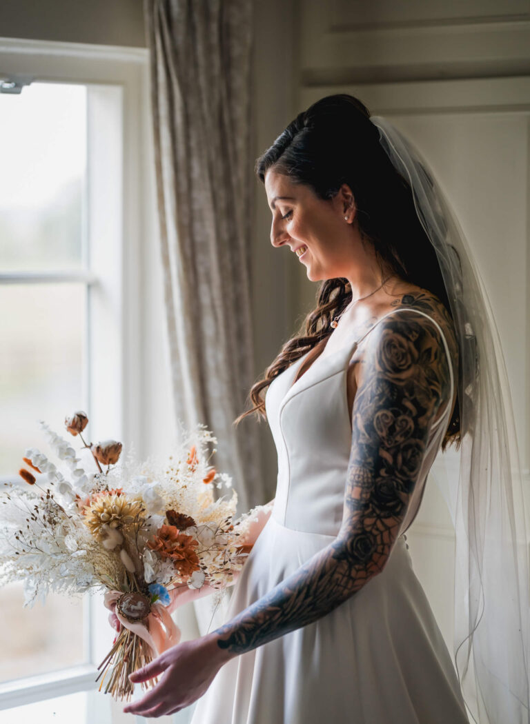 Bride holding bouquet by window, smiling gracefully.