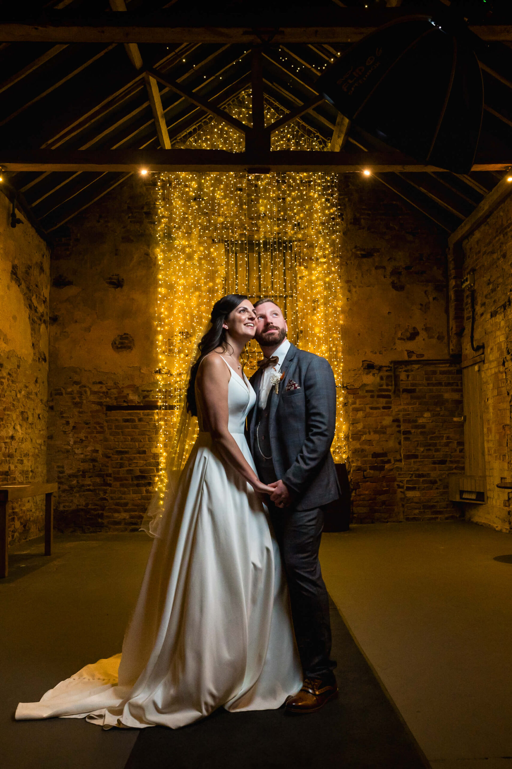 A bride and groom stand together in a rustic venue, holding hands and smiling, with strings of glowing lights cascading behind them.