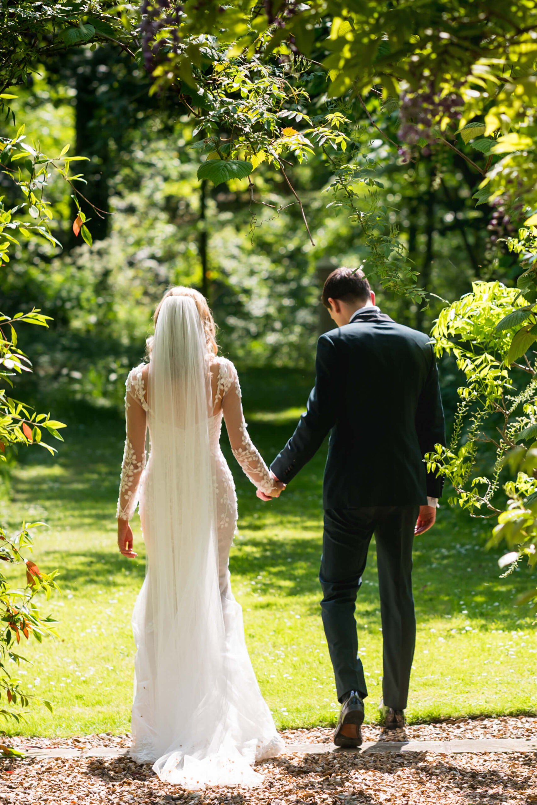 A bride in a long white dress and veil holds hands with a groom in a dark suit as they walk together outdoors in a sunlit garden.