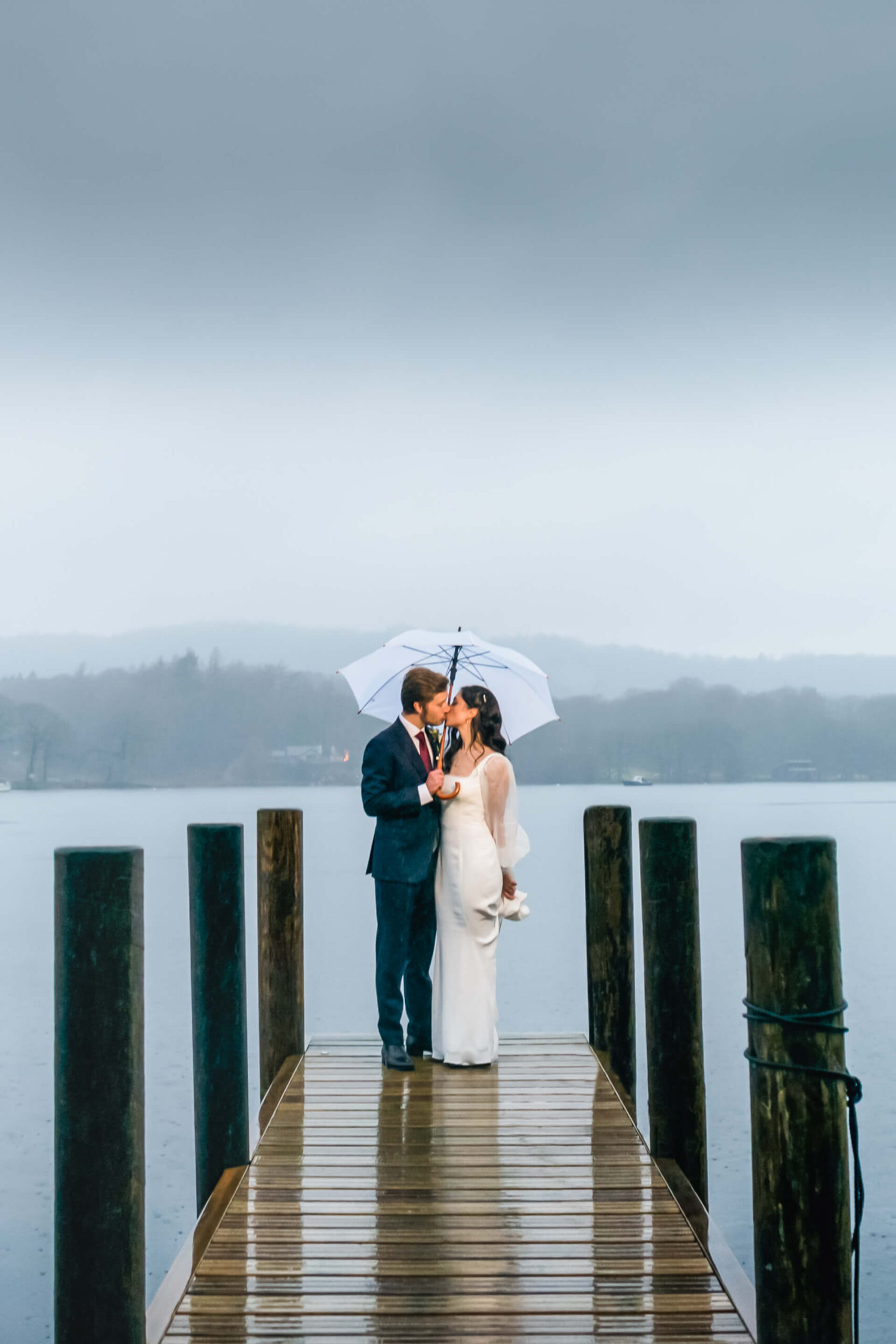 A couple stands on a jetty over a lake, facing each other and sharing a white umbrella on a rainy, overcast day.
