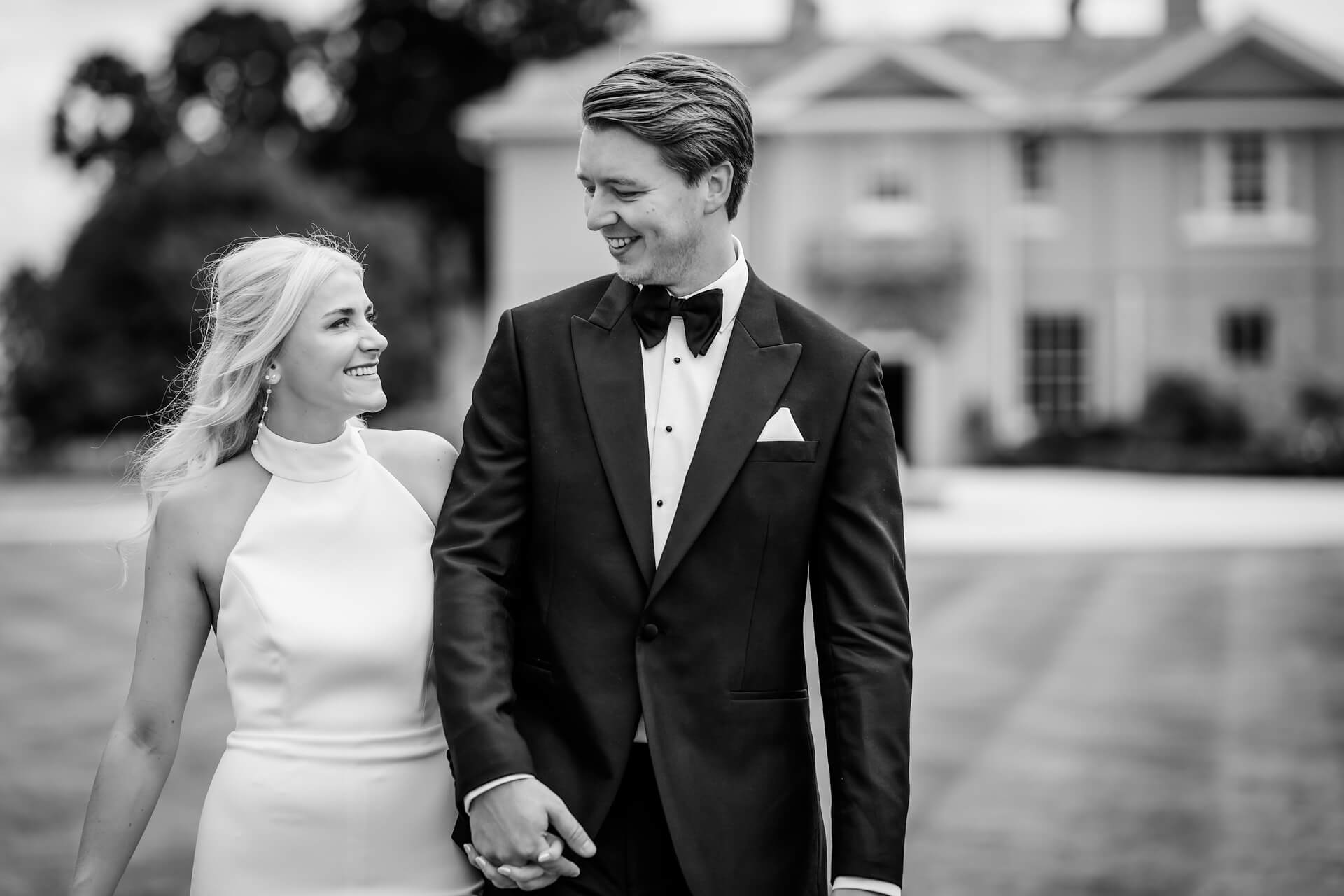 A couple dressed formally, with the woman in a white dress and the man in a black dinner suit, hold hands and smile at each other outside a large house.