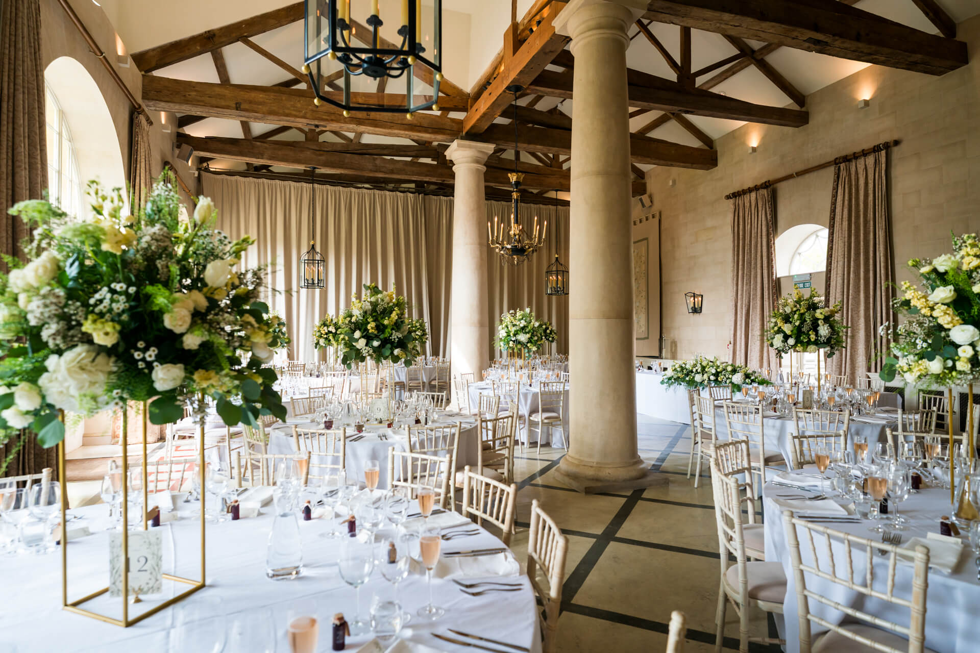 A decorated banquet hall with round tables set for an event, featuring white tablecloths, tall floral centrepieces, and wooden beams on the ceiling.