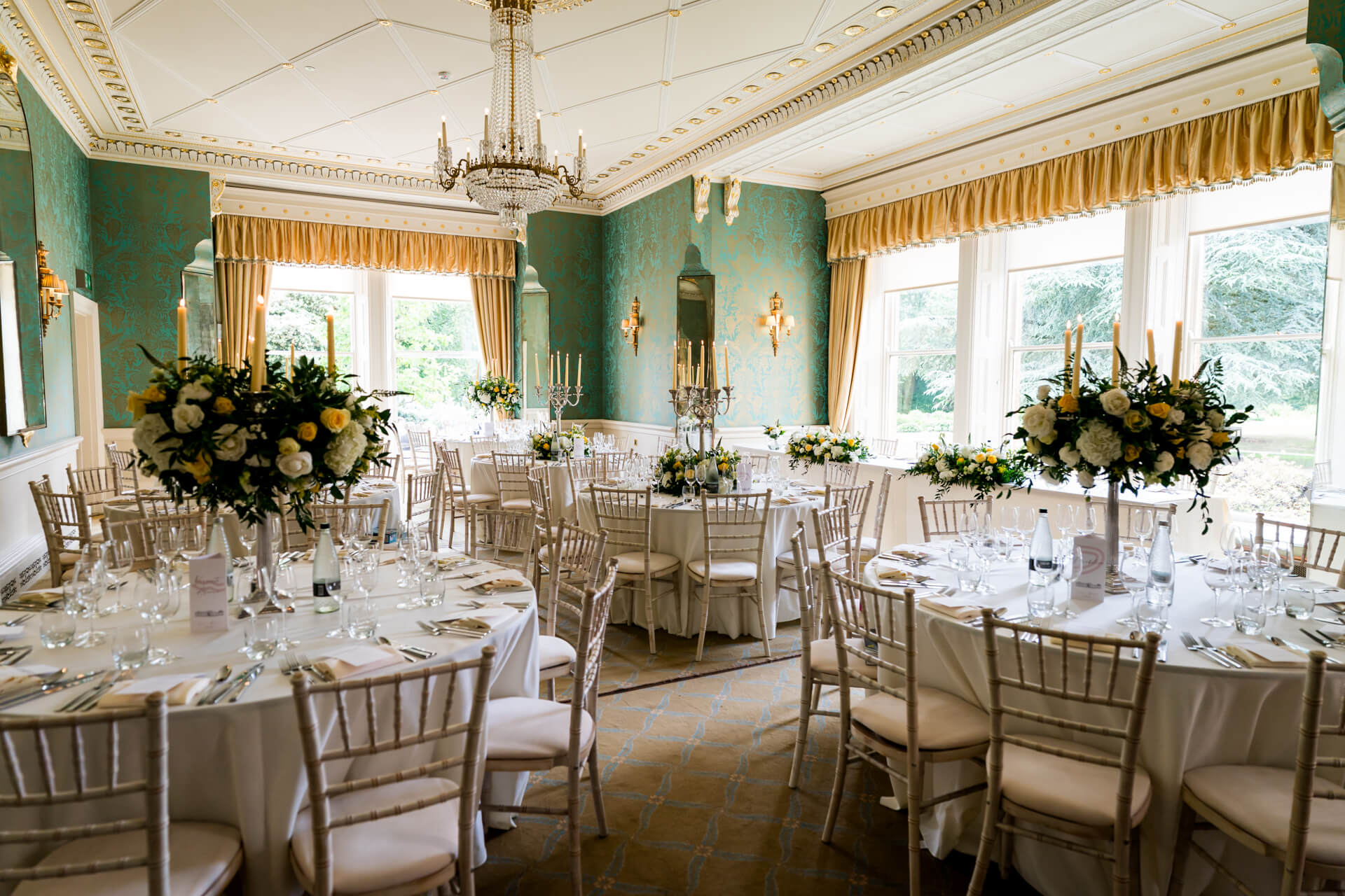 Elegant banquet hall with round tables set for an event, featuring white tablecloths, floral centrepieces, and large windows letting in natural light.