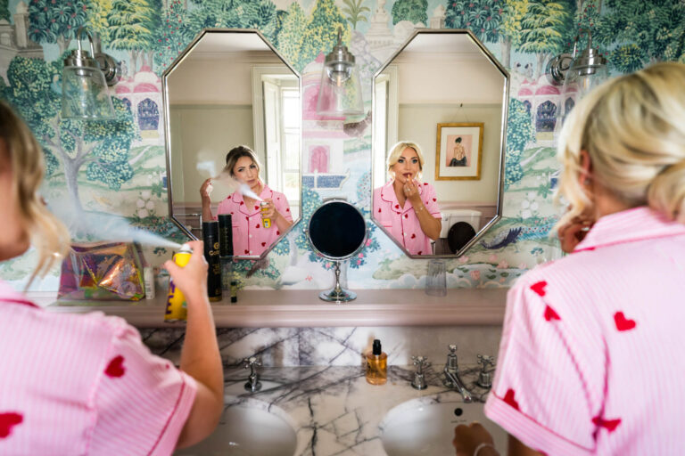 Woman styling hair and applying lipstick in mirror.