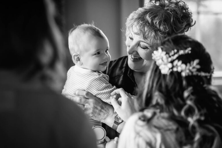 Smiling grandmother holding baby, surrounded by family.