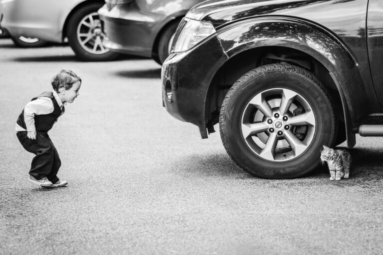 Child curiously observing kitten beside parked car wheel.