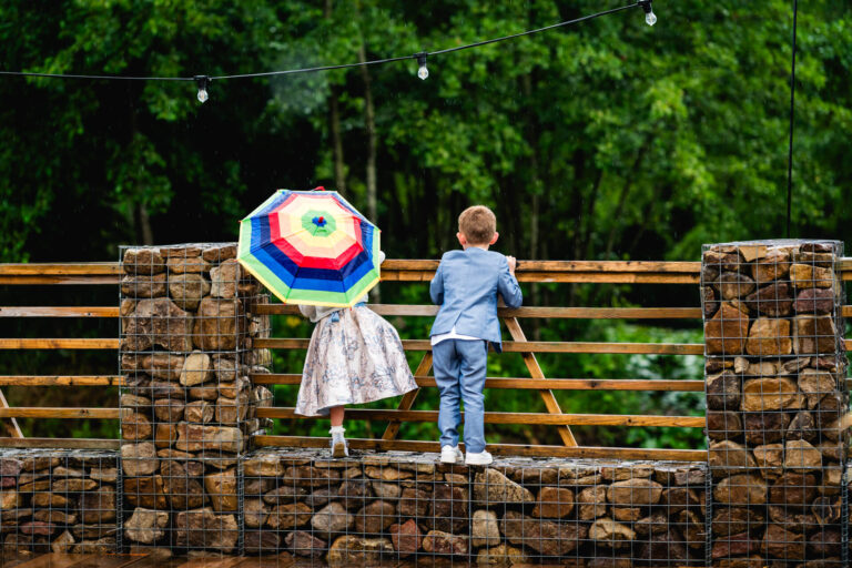 Children on bridge, one with rainbow umbrella, rainy day.